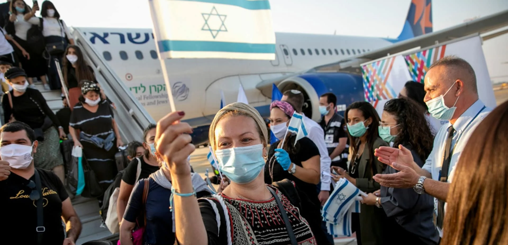 De nouveaux immigrants descendant d’un avion de la Keren Layedidout brandissent des drapeaux israéliens à leur arrivée en Israël, accueillis avec joie sur le tarmac.